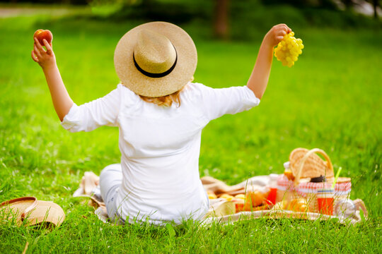 Happy blonde woman in straw hat with peach and grapes on picnic blanket. Raised arms, basket, summer vacation and country freedom concept, rear view from back.