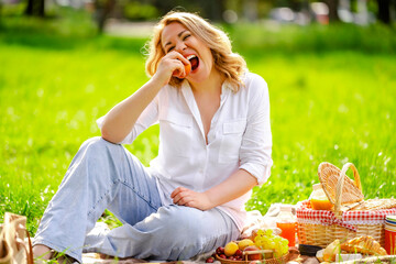 Obraz premium Laughing blonde woman with fresh peach on picnic blanket in park. Cheerful emotions, summer fruit snack and healthy eating concept.