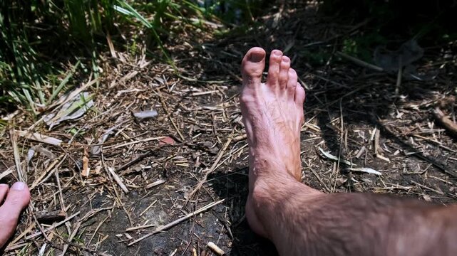 A first-person view shows hairy male legs and bare feet resting on dry leaves and dirt in the woods. Concept of earthing, connecting with nature, and relaxation during a summer hike.