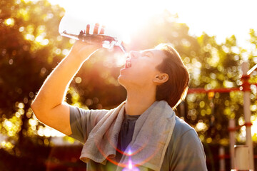 A thirsty man is drinking water outdoors after a fitness workout. Covered in sweat with a towel around his neck, he cools down under the bright sun. © Kyrylo