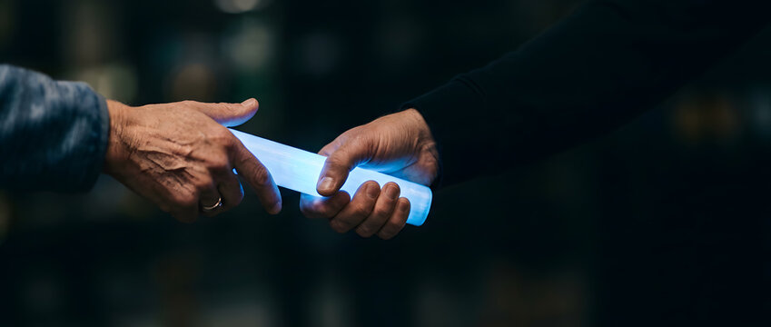 Two hands passing glowing blue cylindrical baton in dark setting representing transition and relay