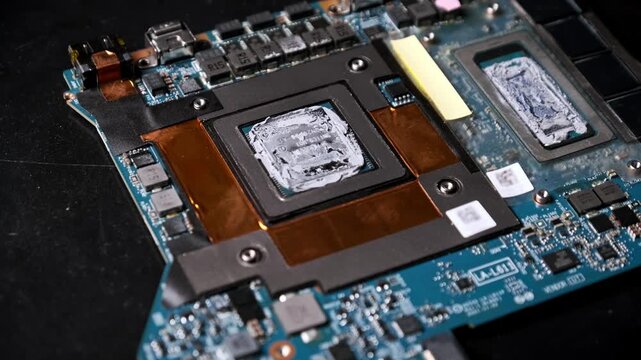An orbiting macro shot captures a gaming laptop graphics processing unit. The chip is covered in thermal paste with the motherboard layout visible in the background.