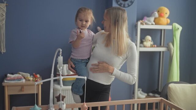 Woman holds toddler on her hip while adjusting a crib mobile and touching the child's hand in a nursery building; maternal tenderness.