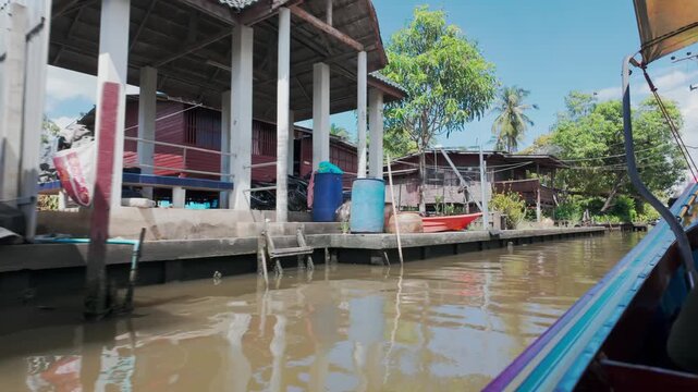 A longtail boat navigates a canal in Bangkok, Thailand, passing by stilted homes and lush vegetation. The scene captures the essence of daily life on the waterways.