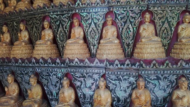 Rows of golden Buddha statues adorn a temple wall in Wat Sam Phran, Nakhon Pathom, Thailand. The intricate mother-of-pearl inlay depicts mythical birds and floral motifs.