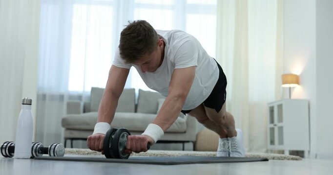 Athletic young man exercising with ab roller on fitness mat at home