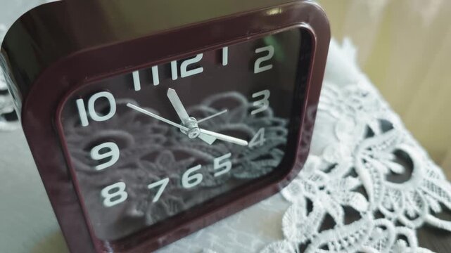 Close-up of a brown vintage-style analog alarm clock resting on a white lace doily. The steady ticking of the second hand evokes nostalgia and the simplicity of daily life.