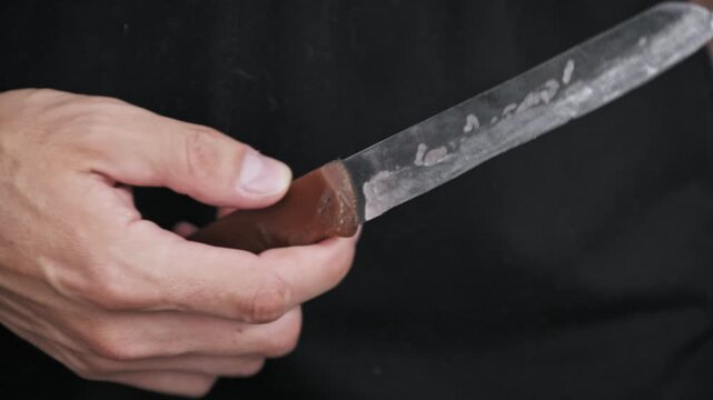 A close-up view of a male hand holding a knife with a wooden handle, turning it to inspect the blade against a dark background. Concept of danger, criminal intent, and weapon possession.