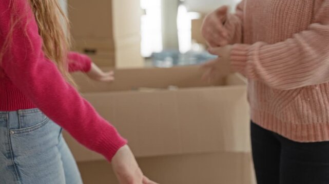 Woman and daughter reaching and passing items with open hands over large cardboard boxes inside building while unpacking and moving; family warmth.