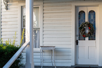 Fototapeta premium The entrance to a historic white building with a double glass window-style door. A colorful, festive Christmas wreath hangs on the vintage door. The entry has a small white table on the porch steps.