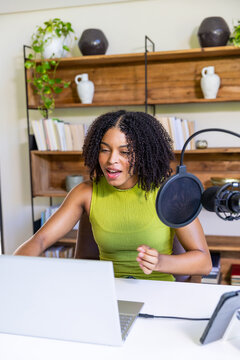 African American woman recording in home studio at desk wearing green top using mic and laptop