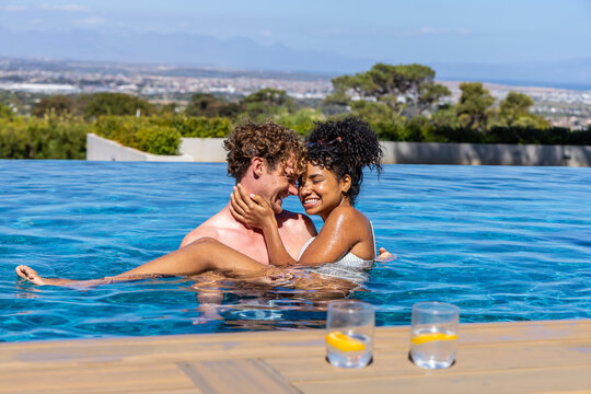 Diverse couple embracing in infinity pool at rooftop wooden deck in swimwear with two lemon glasses