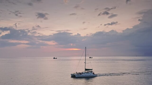 Catamaran carrying passengers across the calm ocean against a beautiful sky with sun setting on the horizon, creating peaceful and relaxing travel experience on Phuket island. Aerial view drone shot