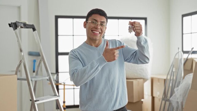Man smiling points finger at key fob while holding keys among moving boxes and ladder in building; new home joy.