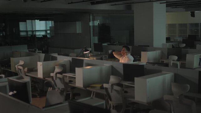 A businessman sitting alone at a dimly lit desk in a dark empty office late at night with computer monitors visible in the background.