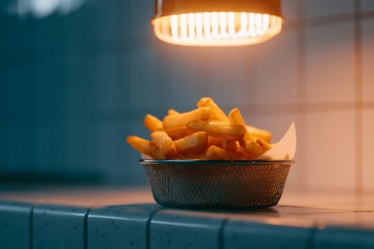Basket of golden fries illuminated under a warm light, placed on a tiled countertop in a diner setting with a blurred background