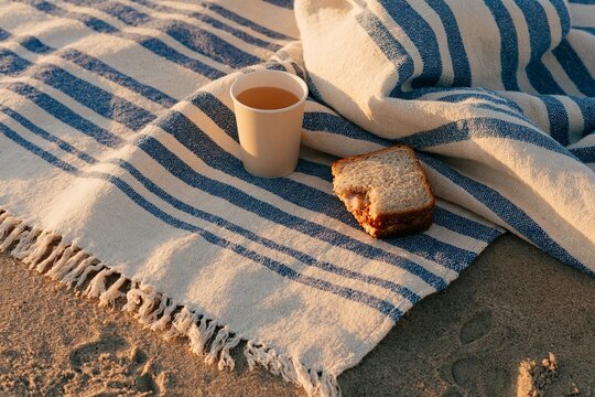 Striped beach towel with a paper cup and a sandwich resting on sandy shore during sunset, creating a serene coastal picnic scene