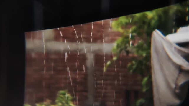 Close-up shot of heavy rain pouring down from a corrugated metal roof against a blurred green tree background. Peaceful yet dramatic rainy season atmosphere, capturing the texture of falling water