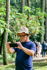 Fototapeta premium Senior Asian Man Taking Photo with His Smartphone While Exploring Lush Green Forest Park
