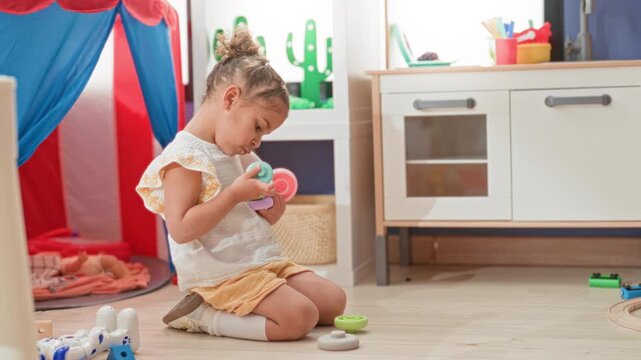 Girl kneeling holds stacking rings toy in playroom, arranging colorful pieces by hand; concentration learning curiosity.