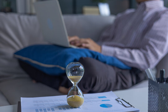Selective focused on hourglass on the table with man working on laptop sitting on sofa at home. Concept for time management and extra over time working.