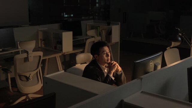 A businessman resting his chin on his fist in a pensive pose while sitting alone at a dimly lit office desk late at night.