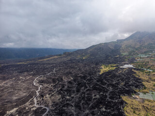 Obraz premium Aerial view of black volcanic lava fields and winding dirt tracks at the foot of Mount Batur under a dramatic cloudy sky in Bali, Indonesia