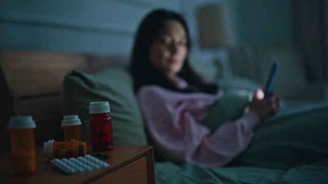Sad brunette resting bed watching cellphone surrounded by medicine bottles