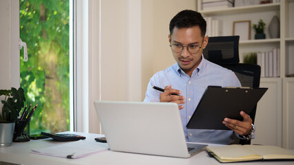 Fototapeta premium Smiling businessman working on laptop with financial documents in a modern home office.