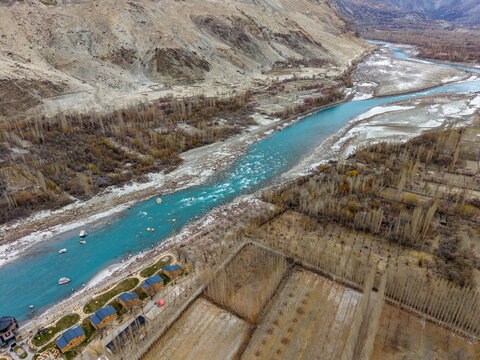 Aerial view of the turquoise Shyok River flowing through the valley with small wooden cabins along the bank, bare trees, and rugged mountain slopes in Ghawari, Gilgit Baltistan, Pakistan.