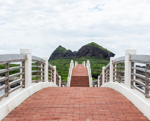 Obraz premium Sanxiantai Arch Bridge in Taiwan. Red brick pathway leads to volcanic rock formations. Lush green hills under a cloudy sky. Coastal walkway view.