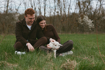 Obraz premium Lifestyle scene with a couple sitting on lush grass outdoors, gentle interaction with a white goat kid, smiling and relaxed, natural light, candid moment of countryside bonding