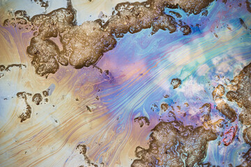Fototapeta premium Iridescent oil slick spreading on a muddy spring puddle with melting snow, showing vivid gasoline stains and toxic chemical pollution pattern on the asphalt road surface