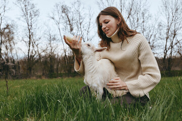 Obraz premium Cheerful woman outdoors, feeding and cuddling a goat kid in a sunlit meadow, warm sweater, gentle interaction, natural colors, relaxed countryside moment