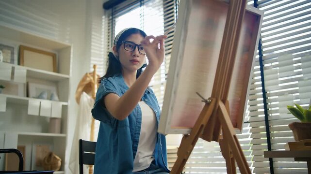 Young Asian woman artist painting on canvas easel with paintbrush in art studio, representing creative professional women designer, fine arts student lifestyle and freelance illustrator