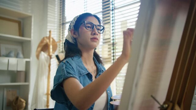 Young Asian woman artist painting on canvas easel with paintbrush in art studio, representing creative professional women designer, fine arts student lifestyle and freelance illustrator