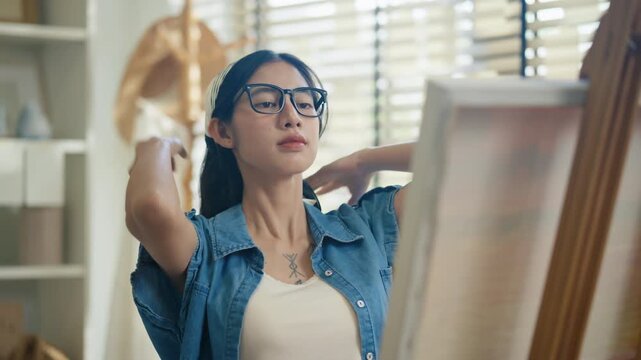 Young Asian woman artist painting on canvas easel with paintbrush in art studio, representing creative professional women designer, fine arts student lifestyle and freelance illustrator