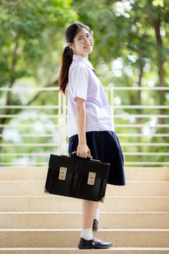 Portrait of a cheerful female student in a traditional Thailand high school uniform, featuring a white short-sleeved shirt with a small bow tie, a navy blue pleated skirt.