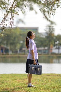 Portrait of a cheerful female student in a traditional Thailand high school uniform, featuring a white short-sleeved shirt with a small bow tie, a navy blue pleated skirt.