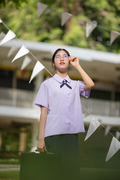 Portrait of a cheerful female student in a traditional Thailand high school uniform, featuring a white short-sleeved shirt with a small bow tie, a navy blue pleated skirt.