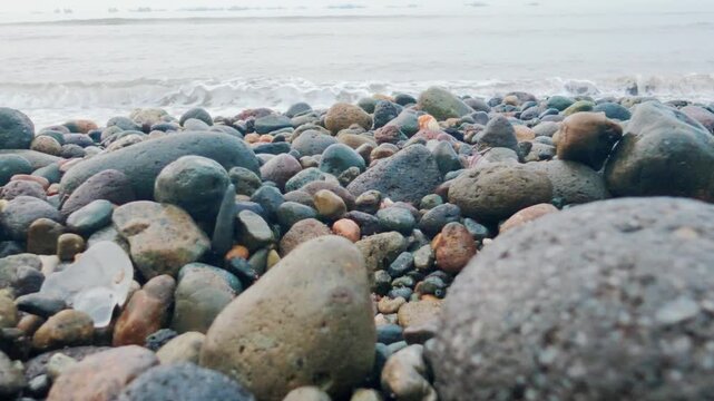 Low-angle shot of gentle ocean waves washing onto a rocky pebble beach with plastic pollution. 