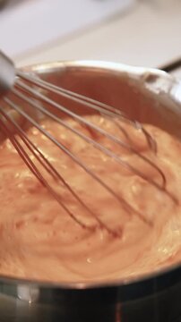 Vertical video. Kitchen work. Cream preparing. Mixing hot frothy red liquid in metal bowl on stove adding milk. Confectionery whisk technique.