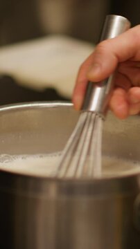 Vertical video. Cooking process. Stirring whisk. Metal saucepan with milk heating on stove. Chef hand mixing white liquid cream ingredients for delicious dish.