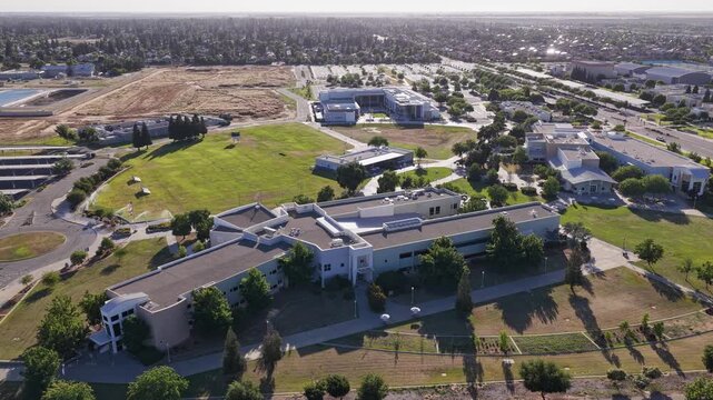 Wide tracking aerial view showing the modern buildings and green landscape of the Clovis Community College campus in Fresno, California, with the surrounding city visible in the background