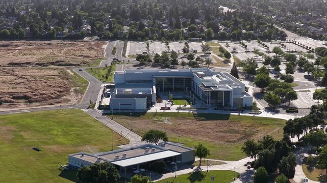 Aerial push out shot of the modern architectural buildings of Clovis Community College campus in Fresno, California, surrounded by green lawns, trees, and a large, almost empty parking lot