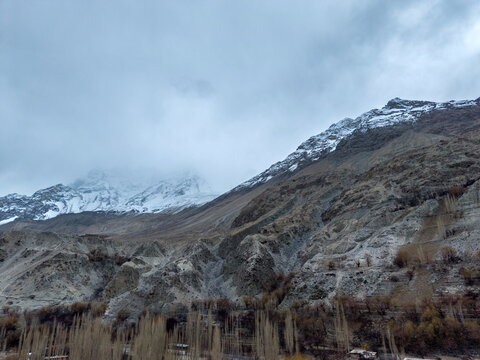 Aerial view of Khaplu valley featuring rugged snow-capped mountains, steep rocky slopes, and rows of bare trees under a cloudy sky in Khaplu, Gilgit Baltistan, Pakistan.