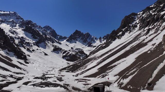 Ascending drone footage revealing snow-covered Andes peaks and the international border corridor connecting Chile and Argentina. The scene includes mountain infrastructure surrounded by dramatic alpin