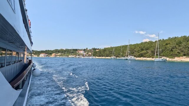 View from a cruise ship departing Fiskardo harbor on Kefalonia Island, Greece. Sailing yachts anchored along the pine-covered coastline while the vessel leaves the marina under clear summer skies