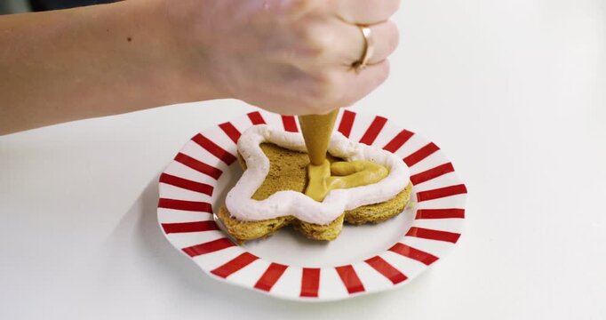 Confectionery process. Cream cookie. Baker hand adding sweet caramel topping mass on gingerbread man with piping bag on striped plate.