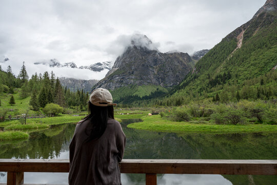 The silhouette of a woman leaning against a wooden railing, gazing out at the mountain lake and the mist-shrouded peaks
